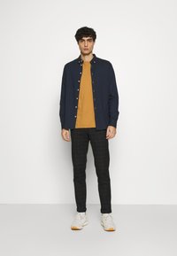 Marineblaues Hemd mit Knöpfen über einem senffarbenen T-Shirt, kombiniert mit schwarzer karierten Hose und weißen Sneakern mit braunen Sohlen.