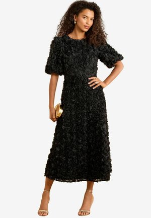 Rochie maxi neagră cu aplicații de trandafiri texturate, mâneci bufante și talie strânsă. Include o poșetă aurie și sandale cu barete.