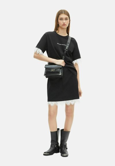 Modelo femenina vestida con un vestido negro con encaje blanco, botas negras hasta el tobillo y carrying una bolsa cruzada negra contra un fondo blanco.