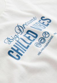 Tissu blanc avec un texte bleu "Big Dreams CHILLED VIBES" et de petites icônes au-dessus, imprimé avec "ISLAND PARADISE - 1982".