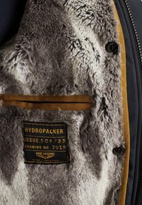 Černá a šedá bunda s kožešinovou podšívkou uvnitř, s nálepkou "HYDROPACKER." Disponuje hladkou texturou a hnědým akcentem na části zipu.