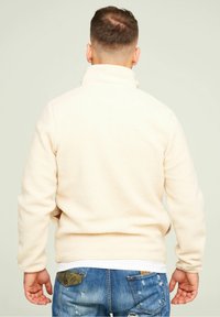 Kremowa bluza polarowa z wysokim kołnierzem, z teksturowaną powierzchnią. W połączeniu z poszarpanymi niebieskimi dżinsami, odsłaniającymi tylne kieszenie.