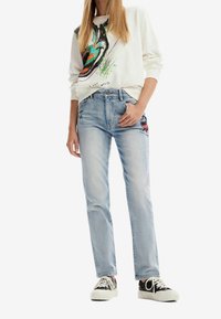 Jeans in denim blu chiaro con gamba dritta, dotati di ricami floreali su una gamba. Abbinati a una felpa beige con stampa grafica colorata.