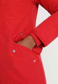 Rote Jacke mit strukturiertem Oberflächen, Druckknopf-Tasche und gerippten Bündchen. Das Design umfasst einen leichten Glanz und glattes Material.