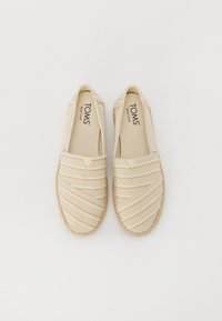 Chaussures slip-on en toile beige avec un motif rayé, semelle en jute et côtés élastiques pour un enfilage facile. Nom de la marque "TOMS" visible sur la semelle intérieure.
