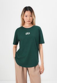 Tamnozelena pamučna majica s kratkim rukavima, okruglim izrezom i bijelim "GAP" logotipom na prsima, u kombinaciji sa svijetlim bež hlačama širokih nogavica.
