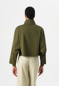 Chaqueta verde oliva recortada con un cuello alto, mangas anchas y una textura suave. Combinada con pantalones beiges claros y vista desde atrás.