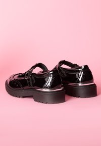 Pantofi din piele lăcuită neagră cu toc masiv, perforații decorative, o curea cu cataramă și o talpă texturată.