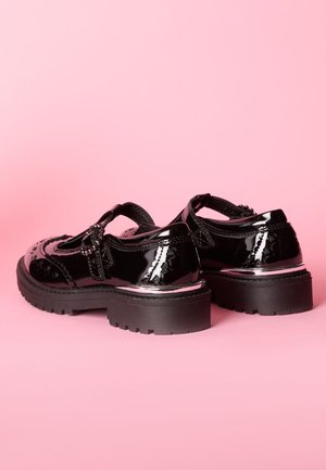 Pantofi din piele lăcuită neagră cu toc masiv, perforații decorative, o curea cu cataramă și o talpă texturată.