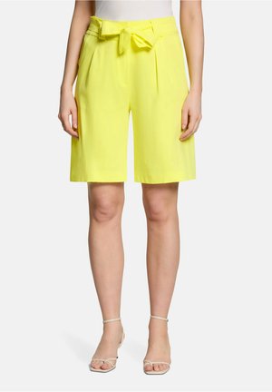 Kobieta nosząca jasne żółte krótkie spodenki do kolan z przednimi zakładkami i wiązanym materiałowym paskiem, w połączeniu z białymi sandałami na obcasie z paseczkami.