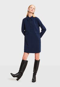 Námornícky modré šaty s golierom, s gombíkovou légou a dvoma prednými vreckami. Nosené s čiernymi koženými čižmami po kolená. Hladký povrch textílie.