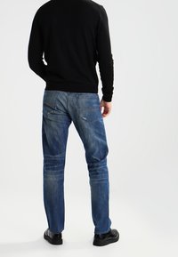 Suéter oscuro combinado con jeans de mezclilla azul claro de corte relajado, con un aspecto desgastado y bolsillos traseros tradicionales. Zapatos negros completan el conjunto.