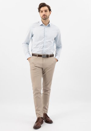 Mężczyzna o ciemnych włosach, ubrany w jasnoniebieską koszulę, beżowe spodnie, brązowy pasek oraz brązowe buty, stojący z rękami w kieszeniach.