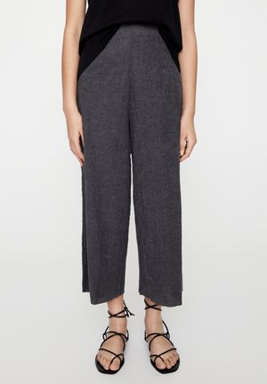 Femme portant un pantalon gris texturé taille haute, jambes larges, associé à des sandales plates noires à lacets et un haut noir, debout sur fond blanc.