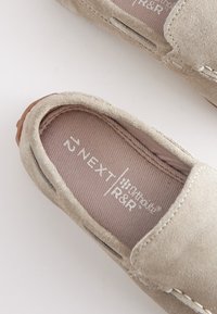 Beige suede slip-on shoes with stitched detailing and a soft fabric lining. Brand logo visible on the insole. Round toe design.