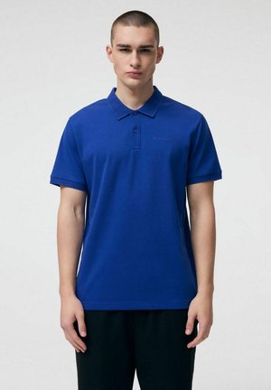 Młody mężczyzna z krótkimi włosami, ubrany w prostą, krótką koszulkę polo w kolorze królewskiego błękitu oraz czarne spodnie, stojący na białym tle.