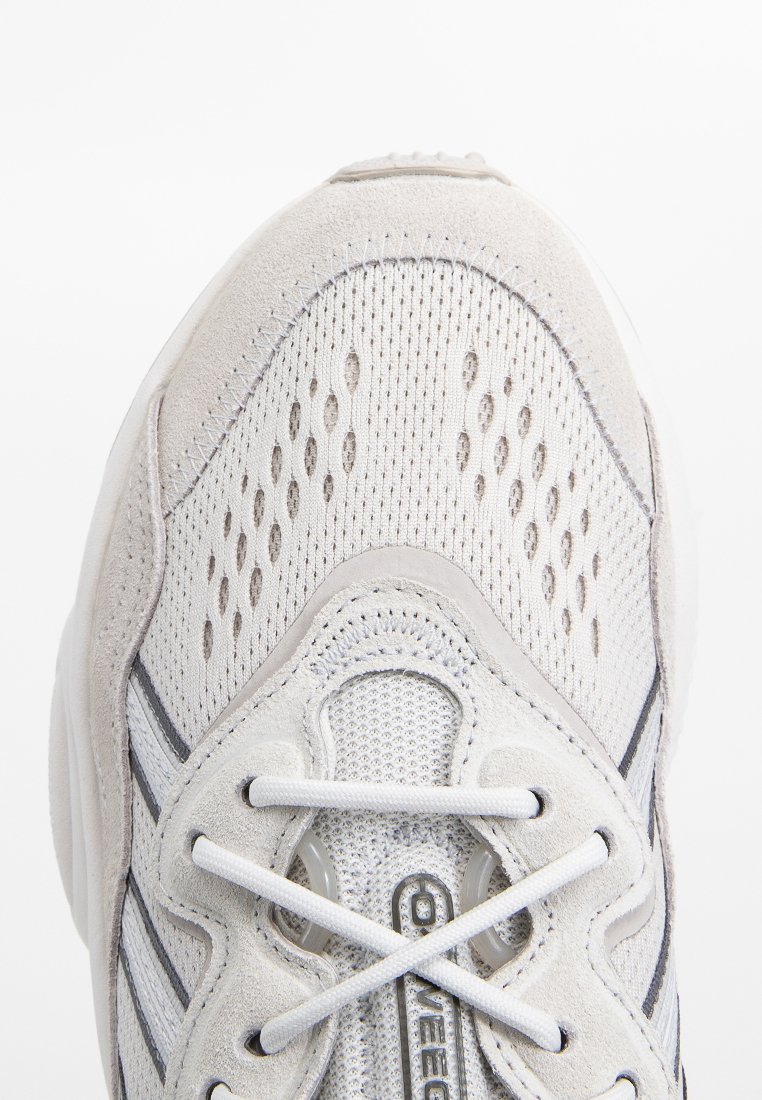 Zapato deportivo blanco con una parte superior de malla y acentos de ante, perforaciones para ventilación y una punta redonda. Cintas blancas y rayas grises.