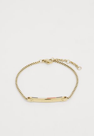 Kulta-tona metallinen ranneke, jossa on joustava käärmehopeaketju. Rannekkeessa on suorakulmainen levy, jossa on "HILFIGER"-kirjoitus ja värilliset aksenttiraitat.