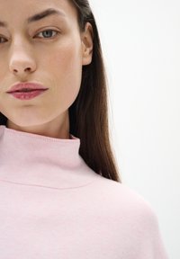 Šviesiai rožinė, raukšlėta golfo megztinė su aukštu apykaklę, minkšta tekstūra, minimalios siūlės ir laisvalaikio siluetas. Matomos odos atspalvio lūpos ir ilgi plaukai.