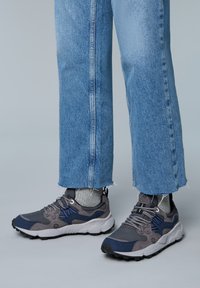 Scarpe sportive grigie e blu navy con un upper texturizzato, dotate di lacci, punta rinforzata e suola bianca a contrasto. Indossate con jeans a pozzetto.