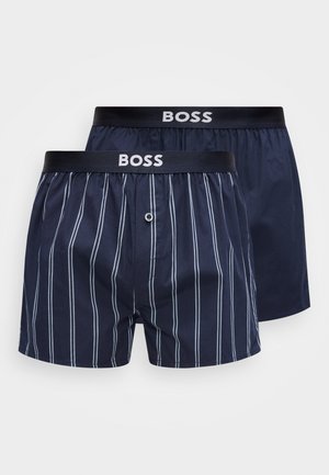 Deux paires de boxers bleu marine avec des ceintures noires portant le mot "BOSS" ; une paire a des rayures blanches verticales, l'autre est unie bleu marine.