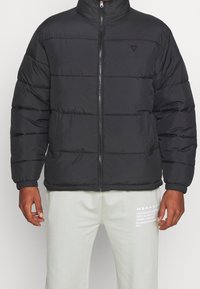 Giacca a piumino nera con collo alto, chiusura frontale con cerniera, design trapuntato orizzontale e polsini elastici. Indossata sopra pantaloni della tuta grigio chiaro.