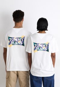 Las camisetas de algodón blancas presentan un gráfico colorido de "TOMMY JEANS" en la parte trasera, rodeado de elementos florales y un marco contrastante.