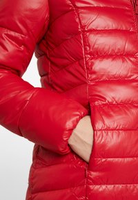 Chaqueta acolchada roja con un exterior brillante, con costuras horizontales estrechas, cierre de cremallera y un bolsillo lateral. Material suave y con aislamiento.