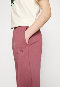 Vaalean värinen t-paita, jossa on vihreä grafiikka, yhdistettynä vaaleanpunaisiin collegehousuihin, joissa on sivutasku ja brodeerattu logo reidessä.