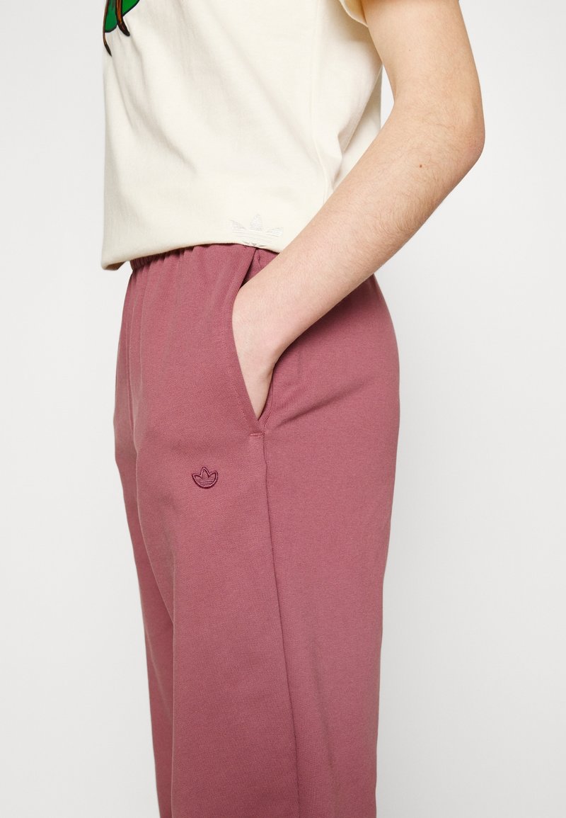 Vaalean värinen t-paita, jossa on vihreä grafiikka, yhdistettynä vaaleanpunaisiin collegehousuihin, joissa on sivutasku ja brodeerattu logo reidessä.