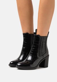 Bottines noires en matériau brillant embossé crocodile. Elles sont dotées de panneaux latéraux élastiques, d'un bout arrondi et d'un talon haut épais pour plus de stabilité.