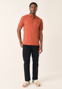 Terracotta gekleurd poloshirt met een kraag en korte mouwen, gecombineerd met zwarte broek. Het model draagt multicolor sneakers.