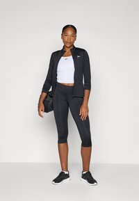 Black athletic jacket, white crop top, black capri leggings, and black sneakers. Model holds a black gym bag. Smooth, fitted materials.