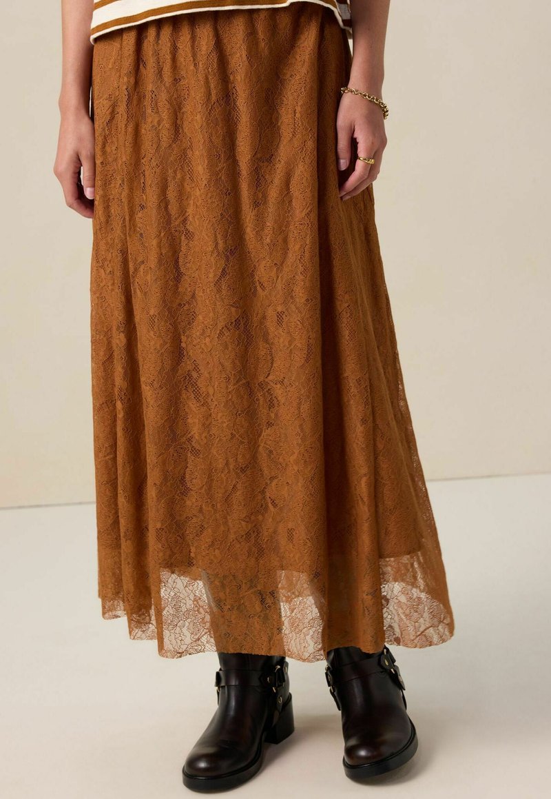 Femme portant une longue jupe en dentelle marron assortie à des bottes noires à la cheville, avec un bracelet et une bague en or visibles sur sa main droite.