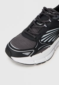 Shoe atlético cinza com parte superior em malha, acentos pretos e detalhes refletivos. Sola acolchoada com padrão de relevos texturizados.