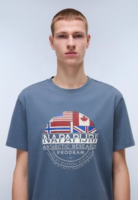 T-shirt en coton bleu avec une impression graphique de drapeaux nationaux et le texte "Programme de recherche antarctique Napapijri" dans des couleurs contrastées.