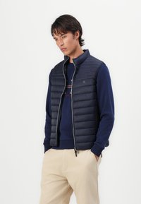 Vesta matlasată de culoare bleumarin, cu guler în picioare, detalii cu fermoar gri și un design ușor, purtată peste un pulover bleumarin și pantaloni bej.