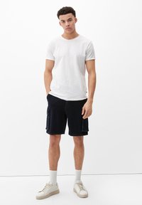Weißes Kurzarm-T-Shirt, dunkle Denim-Cargo-Shorts mit Seitentaschen und beiger Sneaker mit weißen Socken, stehend vor einem einfarbigen Hintergrund.