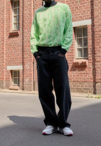 Maglione verde neon con un motivo testurizzato, cinturino nero, abbinato a jeans neri larghi e sneakers bianche con dettagli rossi.