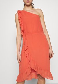 Koralowa sukienka na jedno ramię, z falbaną wzdłuż boku i miękkim, zwiewnym materiałem. Średniej długości, dopasowana w talii.