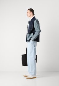 Dongerijakke med mørke og lyseblå sektioner, enkelt brystlomme, kombinert med lyseblå bukser og hvite joggesko, holder en svart tote bag.