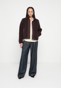 Weinroter, flauschiger Mantel mit hohem Kragen und Reißverschluss vorne, kombiniert mit einem hellgelben Oberteil und weit geschnittenen dunkelblauen Jeanshosen.