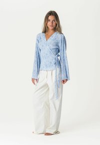 Helle blaue Wickelbluse mit langen Ärmeln, dezentem Blumenmuster und einem Bindeband in der Taille; kombiniert mit weiten weißen Hosen.