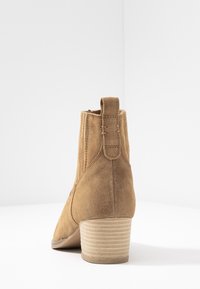 Brązowy zamszowy botek na kostkę z zaostrzonym noskiem, fakturowaną podeszwą i nałożonym obcasem. Posiada pętelkę z tyłu oraz przeszycia dekoracyjne.