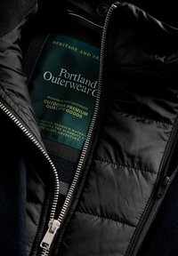 Črno zunanjo jakno z zadrgo spredaj, s prešito notranjo podlogo, mornarsko modrim flisnim zunanjim delom in oznakami, ki pravijo "Portland Outerwear Co."