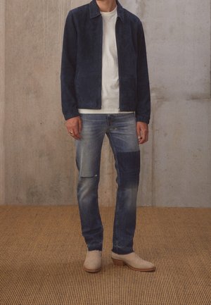 Homme portant une veste zippée bleu foncé, un t-shirt blanc, un jean bleu délavé avec des patchs et des bottes en suède beige, debout sur un sol texturé.