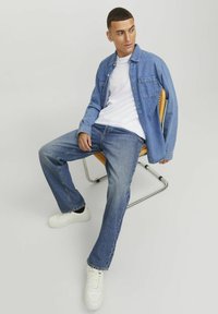 Jeanshemd in Hellblau, weißes T-Shirt, Blue Jeans und weiße Sneakers. Modell sitzt auf einem Metallstuhl und zeigt einen lässigen Schnitt und Casual-Stil.