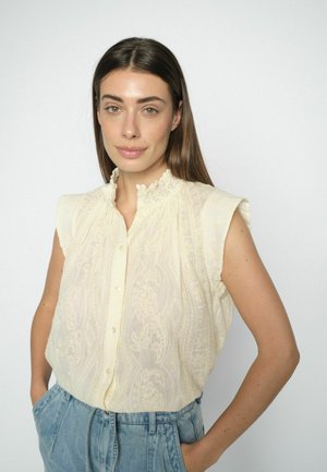 Frau mit langem braunem Haar, die eine ärmellose cremefarbene Spitzenbluse und hoch taillierte hellblaue Jeans trägt, steht mit den Händen in den Taschen vor weißem Hintergrund.