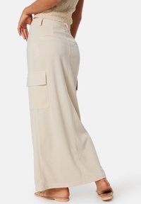 Pantalon beige à jambes larges avec une texture lisse, doté de deux poches cargo sur les côtés et d'une taille normale. Le design est minimaliste.