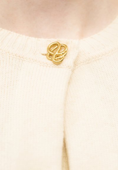 Χρυσή αφηρημένη καρφίτσα σε σχήμα κύκλου που κλείνει το γιακά μιας εκρού πλεκτής μπλούζας πάνω σε γυμνό δέρμα.